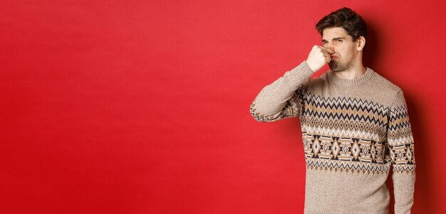 Bild eines angewiderten jungen Mannes im Winterpullover mit geschlossener Nase und Grimassen von schlechtem Geruch, der über rotem Hintergrund steht