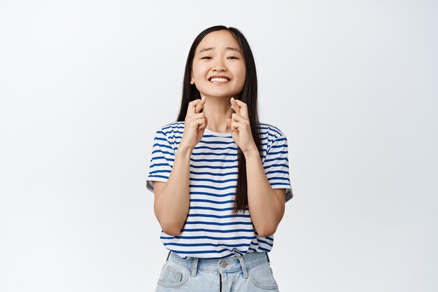 Bild eines asiatischen Mädchens, das lächelt und die Daumen drückt, in der Hoffnung, etwas zu erreichen, das ein positives Ergebnis vorwegnimmt und einen Wunsch macht, der über weißem Hintergrund steht