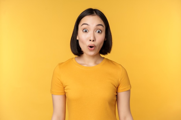 Bild eines asiatischen Mädchens, das überrascht aussieht und überrascht reagiert, die Augenbrauen hochzieht, beeindruckt, wie es vor gelbem Hintergrund steht