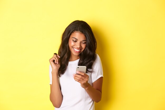 Bild eines attraktiven afroamerikanischen Mädchens in weißem T-Shirt-Messaging auf dem Smartphone, das auf das Handy schaut