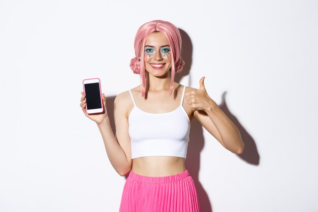 Bild eines attraktiven jungen Partymädchens in rosa Perücke, mit hellem Make-up, Daumen hoch und Handy-Bildschirm, App empfehlen, auf weißem Hintergrund stehend