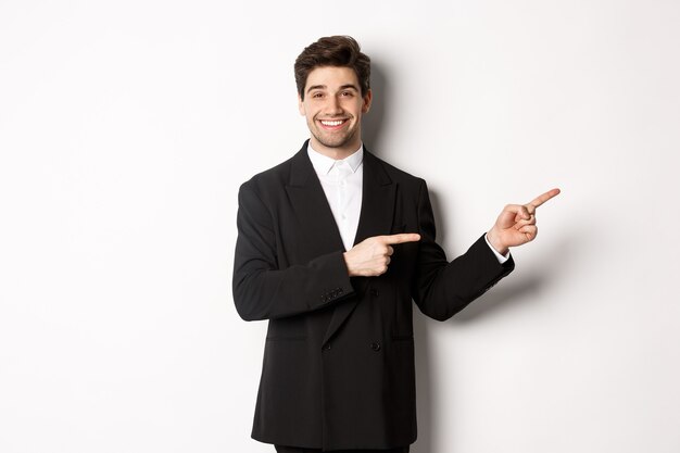 Bild eines attraktiven lächelnden Kerls, der für die Neujahrsparty gekleidet ist, mit den Fingern nach rechts zeigt und Werbung zeigt, die auf weißem Hintergrund steht.