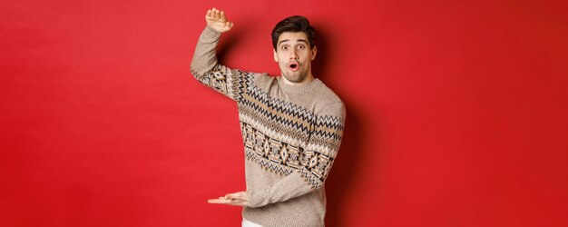 Bild eines attraktiven Mannes im Weihnachtspullover, der ein großes Neujahrsgeschenk formt, etwas Großes und Erstaunliches zeigt und über rotem Hintergrund steht.