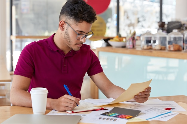 Bild eines beschäftigten unrasierten Texters oder Studenten, der in Freizeitkleidung gekleidet ist, macht Notizen im Notizbuch, fokussiert in Dokument, schaut aufmerksam, posiert in der kleinen Cafetiera, trinkt heißes Getränk.