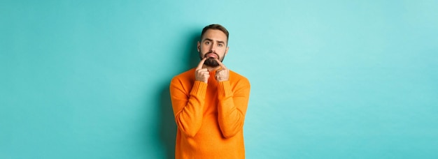 Kostenloses Foto bild eines düsteren bärtigen mannes, der ein trauriges gesicht macht und die stirn runzelt, als er verärgert in einem orangefarbenen pullover gegen tu steht