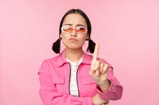 Bild eines ernsthaften, stilvollen asiatischen Mädchens mit Sonnenbrille, das ein Tabu-Schild mit der Aufschrift „Stopp verbieten“ zeigt, das besagt, dass kein Stehen auf rosafarbenem Hintergrund steht