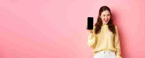 Kostenloses Foto bild eines frechen, attraktiven mädchens, das ihnen den bildschirm des smartphones zeigt, der blinzelt und lächelt und ein handy empfiehlt