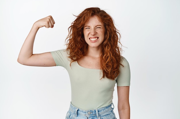 Bild eines frechen rothaarigen Teenagers, der Bizeps beugt, Muskeln am Arm zeigt und glücklich aussieht, Mädchen-Power-Geste, die sich stark vor weißem Hintergrund fühlt