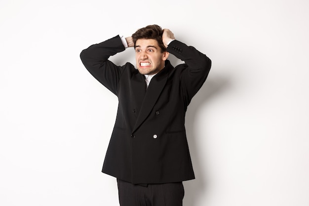 Bild eines frustrierten und wütenden Geschäftsmannes im schwarzen Anzug, der sich die Haare am Kopf reißt und das Gesicht verzieht, nach links in die Katastrophe schaut und vor weißem Hintergrund angespannt steht