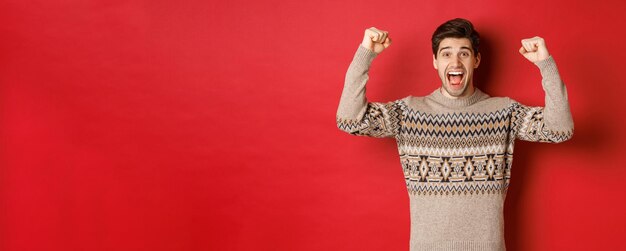 Bild eines glücklichen gutaussehenden Kerls, der sich an Heiligabend freut, die Hände hochhebt und vor Freude schreit, ja, das Neujahrsziel erreicht, das über rotem Hintergrund im Pullover steht