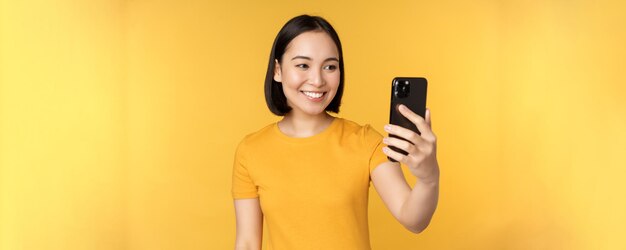 Bild eines glücklichen, schönen asiatischen Mädchens, das auf einer Smartphone-Anwendung spricht, die vor gelbem Hintergrund steht