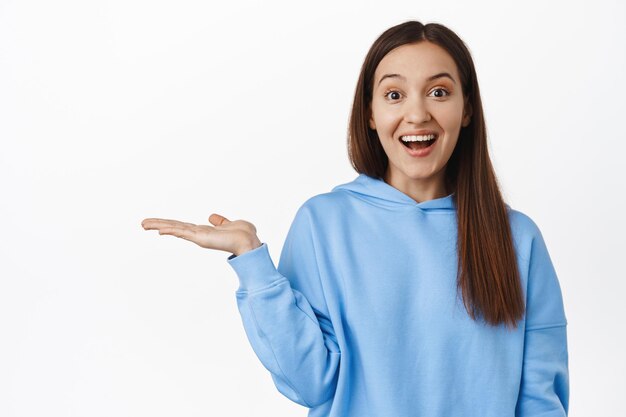 Bild eines glücklichen süßen Mädchens, das in offener Hand hält, aufgeregt über das zum Verkauf stehende Produkt lächelt, Artikel auf ihrer Handfläche zeigt, leerer Raum, vor weißem Hintergrund steht