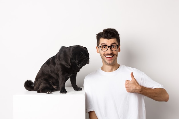 Bild eines glücklichen und zufriedenen jungen Mannes, der in der Nähe von Mopshund sitzt und Daumen hoch zeigt, lächelt und gutes Produkt lobt, weißer Hintergrund