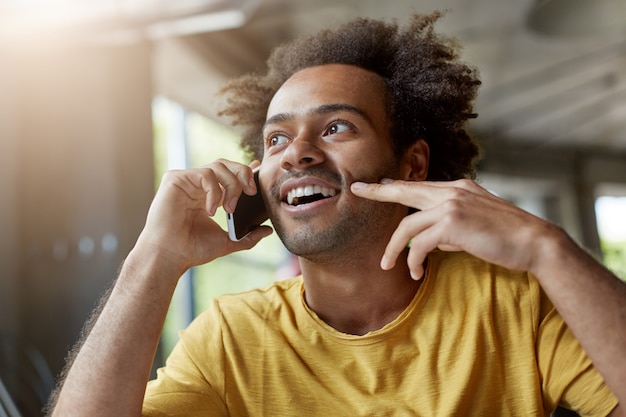 Bild eines gut aussehenden glücklichen afrikanischen Mannes mit Bart und lockigem Haar, der fröhlich lächelt, während er auf Handy spricht, interessierten Blick hat