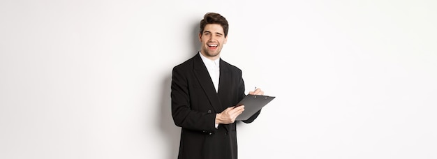 Bild eines gutaussehenden erfolgreichen Chefs im schwarzen Anzug, der blinzelt und lächelt, während er Dokumente im Stehen unterschreibt