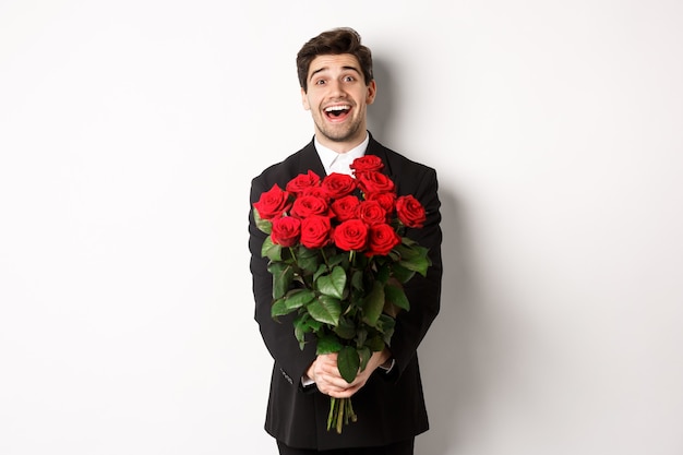 Bild eines gutaussehenden Freundes im schwarzen Anzug, der einen Strauß roter Rosen hält und lächelt, ein Date hat und auf weißem Hintergrund steht