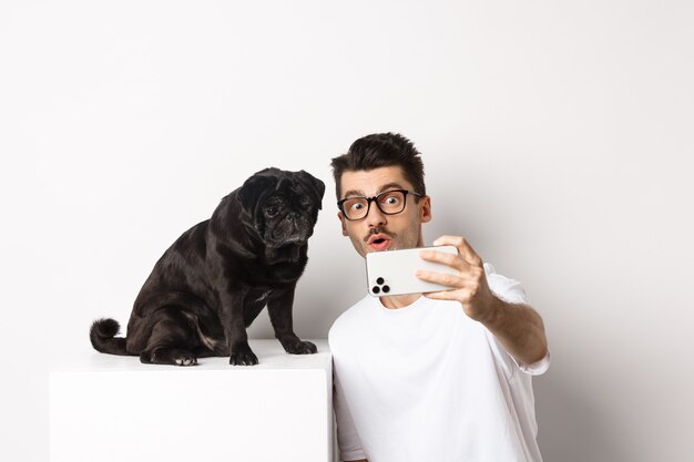 Bild eines gutaussehenden jungen Mannes, der ein Selfie mit einem süßen schwarzen Hund auf dem Smartphone macht und mit Mops auf weißem Hintergrund posiert