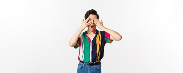 Bild eines gutaussehenden jungen schwulen Mannes mit offenen Augen, um Überraschung zu sehen, die durch Finger späht, mit erstaunten Emotionen, die über weißem Hintergrund stehen