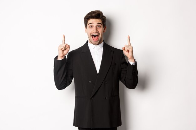 Bild eines gutaussehenden Mannes im Partyanzug, der Ferienpromo zeigt, mit den Fingern nach oben zeigt und erstaunt lächelt, auf weißem Hintergrund stehend