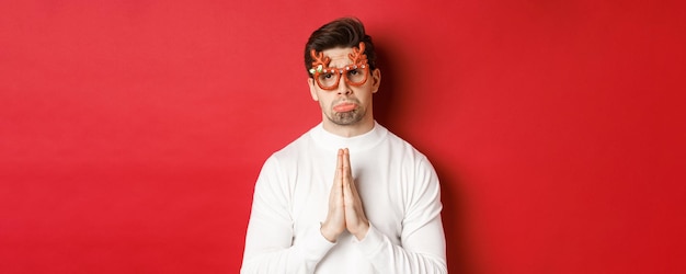 Bild eines gutaussehenden Mannes in Weihnachtsfeierbrille, der um Hilfe bettelt oder sich entschuldigt, einen Gefallen braucht und auf rotem Hintergrund steht