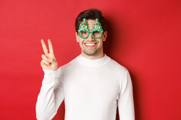 Bild eines gutaussehenden Mannes in weißem Pullover und Partybrille, der ein Friedenszeichen zeigt und lächelt, frohe Weihnachten wünscht und auf rotem Hintergrund steht.