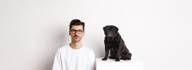 Kostenloses Foto bild eines hübschen hipsters mit brille, der neben einem schwarzen süßen mops sitzt und beide in die kamera starrt