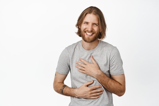 Bild eines kaukasischen, gutaussehenden blonden Kerls, bärtiger Mann, der sorglos lacht und lächelt, glückliches Gesicht eines jungen männlichen Modells in grauem T-Shirt, weißer Hintergrund.