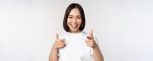 Bild eines lächelnden asiatischen Mädchens, das mit dem Finger auf die Kamera zeigt und sich dafür entscheidet, Sie einzuladen, im T-Shirt vor weißem Hintergrund zu gratulieren