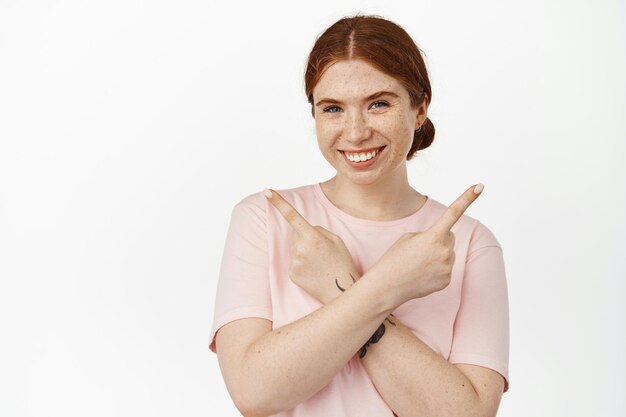 Bild eines lächelnden rothaarigen Mädchens, das mit verschränkten Armen zur Seite zeigt, linke und rechte Produktangebote, Ladenangebote zeigt, glücklich aussieht und vor weißem Hintergrund steht.