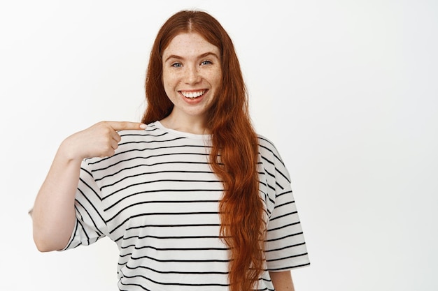 Bild eines lächelnden rothaarigen Mädchens mit langen natürlichen Haaren, das mit erfreutem Gesichtsausdruck auf sich selbst zeigt, weiße Zähne, etwas an ihr zeigt und vor weißem Hintergrund steht.