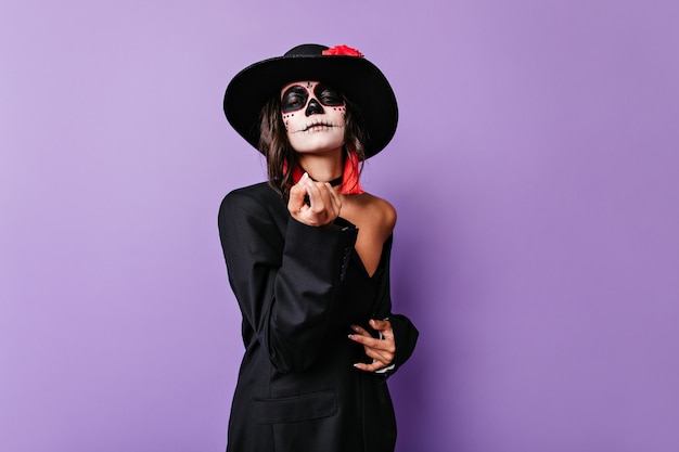 Bild eines Mädchens mit breitkrempigem schwarzen Hut, das zu sich selbst einlädt. Mexikanisches Modell mit Totenkopf-Make-up in übergroßer Jacke.