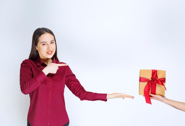 Bild eines Modells der jungen Frau, das auf ein Geschenk mit Bogen auf weißer Wand zeigt.