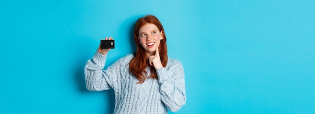Kostenloses Foto bild eines nachdenklichen rothaarigen mädchens, das über das einkaufen nachdenkt und dabei eine kreditkarte vorzeigt und über das stehen nachdenkt