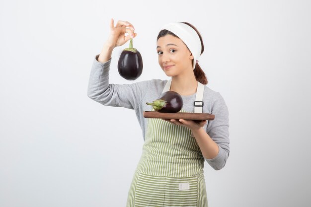 Bild eines netten jungen Frauenmodells in der Schürze, das eine Aubergine hält