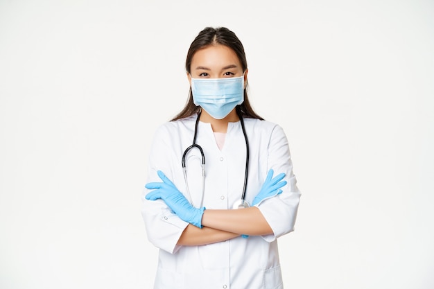 Bild eines professionellen Arztes, asiatische Ärztin in medizinischer Gesichtsmaske, Gummihandschuhe, stehend mit verschränkten Armen, weißer Hintergrund.