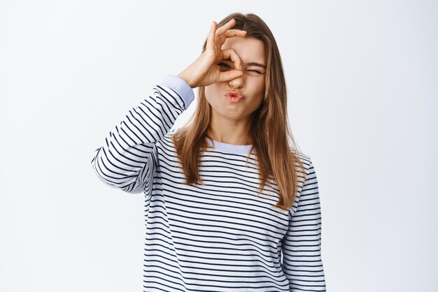 Bild eines stilvollen, attraktiven blonden Mädchens, das ein OK-Zeichen auf dem Auge zeigt, das durch eine Okay-Geste schaut, genehmigt und stimmt zu, sagen Sie in Ordnung, wie es vor weißem Hintergrund steht