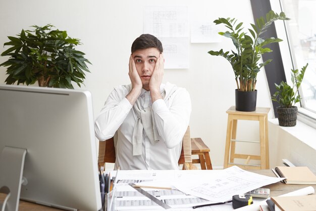 Bild eines verwirrten schockierten jungen männlichen Designers oder Architekten, der im Büro arbeitet, sich gestresst und nervös fühlt, die Hände auf dem Kopf hält, auf den Computerbildschirm starrt und Fehler in seinen Zeichnungen bemerkt