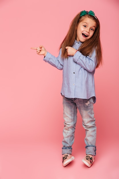 Bild in voller Länge des lustigen kleinen Mädchens, das Zeigefinger-Kopienraum für Ihren Text oder Produkt zeigend gestikuliert