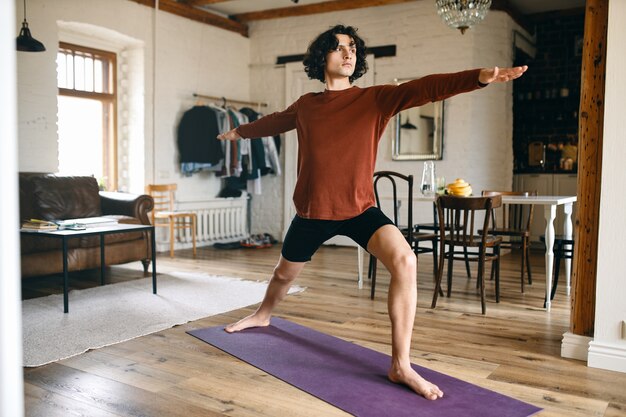 Bild in voller Länge eines gutaussehenden jungen Mannes mit starkem athletischem Körper, der drinnen Yoga praktiziert, in Krieger-2-Pose oder Virabhadrasana steht und tief durchatmet.