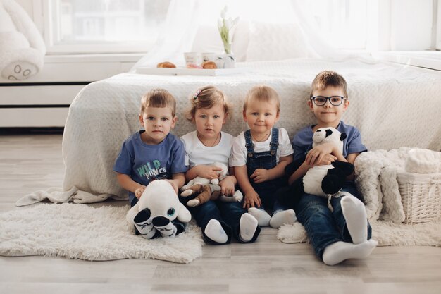 Bild von vier kleinen Kindern in Hauskleidung sitzt in der Nähe des weißen Sofas mit Spielzeug, lächeln und haben Spaß