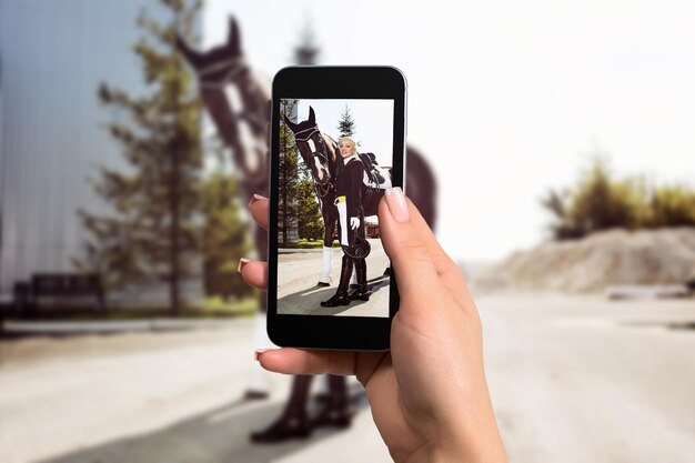 Bild von weiblichen Händen, die ein Mobiltelefon mit Fotokameramodus auf dem Bildschirm halten. Bild einer Reiterin mit einem Pferd.