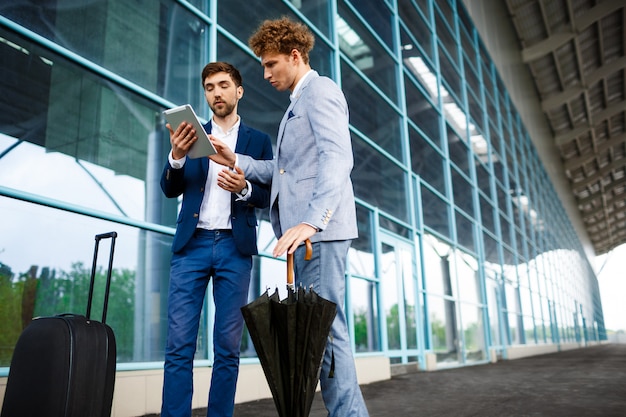 Bild von zwei jungen Geschäftsleuten, die am Flughafen sprechen und Tablette halten
