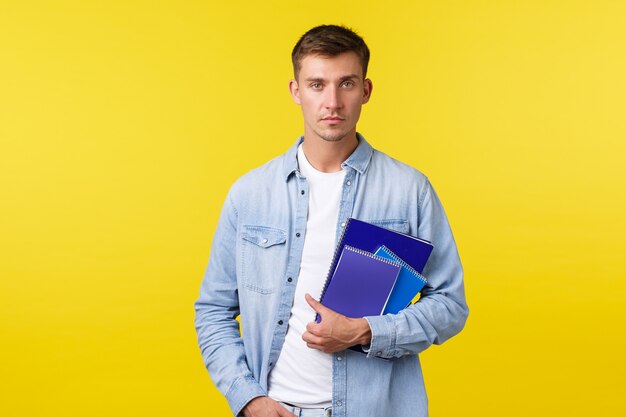 Bildung, Kurse und Hochschulkonzept. Ernsthaft aussehender College-Student, Typ mit Notebooks, der entschlossene Kamera sucht, beiläufiger Ausdruck als Richtung zum Unterricht, gelber Hintergrund.