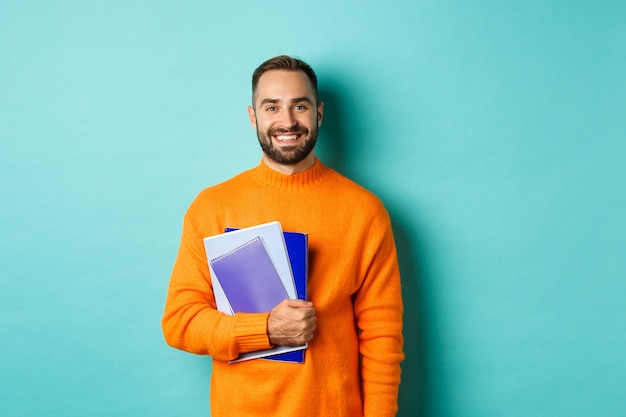 Bildung. Lächelnder bärtiger Mann, der Notizbücher hält und lächelt, Kurse geht, über heller türkisfarbener Wand stehend