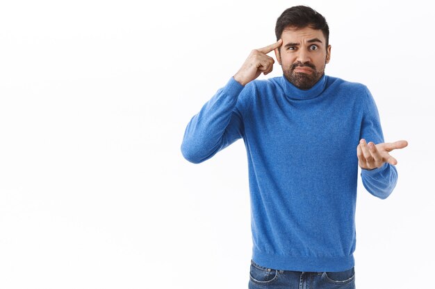 Bist du dumm oder verrückt. Verärgerter und belästigter, frustrierter bärtiger Mann, der Person ausschimpft, weil sie dumme Dinge sagt, sich seltsam verhält, Finger an Schläfe rollen und bestürzt die Hand heben, weiße Wand,