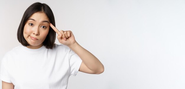 Bist du eine verrückte junge asiatische Frau, die mit dem Finger auf den Kopf rollt und jemanden verspottet, der dumm im T-Shirt über w steht?