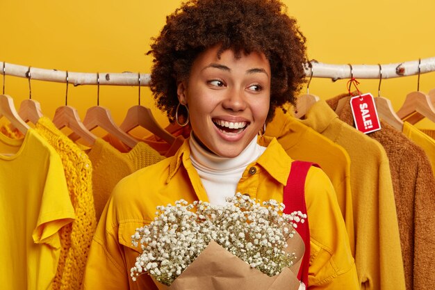 Black Friday und Preissenkungskonzept. Positive lockige Frau freut sich Shop fünfzig Prozent Rabatt auf Angebot, kann viele Outfits für nicht viel Geld kaufen, steht in der Nähe von Vitrine mit gelben Kleidern, trägt Blumen