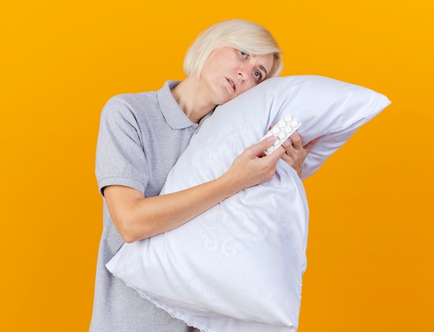 Blasse junge blonde kranke Frau legt Kopf auf Kissen hält Packung von medizinischen Pillen isoliert auf orange Wand