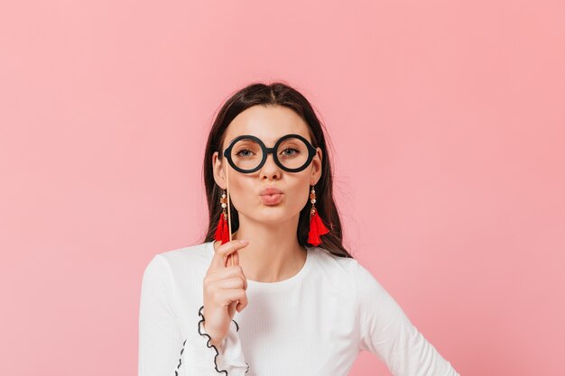 Blauäugige Brünette mit Brille, die auf rosa Hintergrund aufwirft. Dame in weißer Strickbluse bläst Kuss.