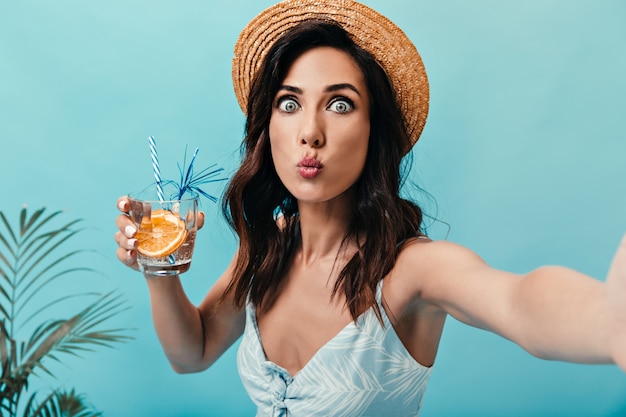 Blauäugige dame pfeift, macht selfie und hält ein glas wasser mit orange